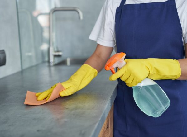 Hände in gelben Handschuhen reinigen eine Küchenarbeitsfläche mit Sprühflasche und Tuch